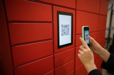 A woman scans a red code to pick up a parcel at a parcel machine. Automated Stock Photos