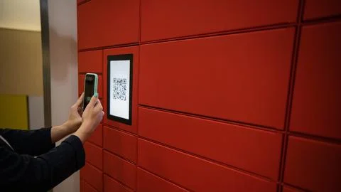 A woman scans a red code to pick up a parcel at a parcel machine. Automated Stock Photos