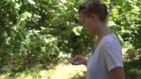Woman scrolls touch screen of her cell phone while walking in a park. Handheld 스톡 동영상 81884835