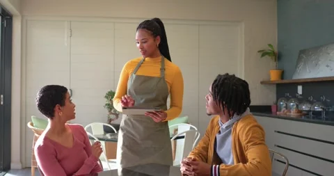 Woman server in apron approaching cafe table greeting couple, presenting menu 動画素材 336174243