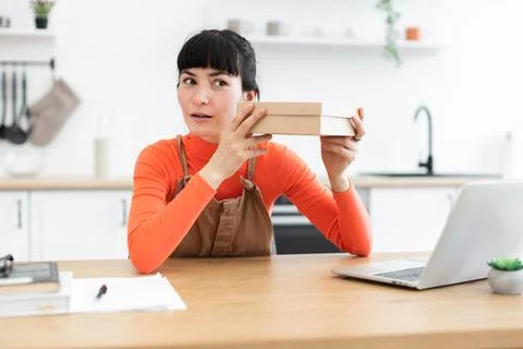 Woman shaking gift box while sitting at kitchen table Stock Photos