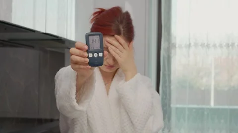 Woman shows glucometer reading of high blood sugar in kitchen setting Stock Footage 346704206