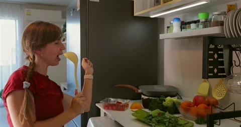 Woman singing with spoon in kitchen, surrounded by fruits and Video stock 305933875