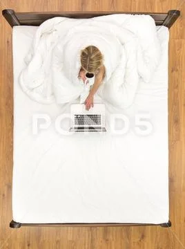 The woman sit on the bed and drink coffee near the laptop. View from above Stock Photos
