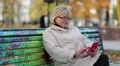 Woman sits on the bench in autumnal city park and uses red smartphone HD Footage