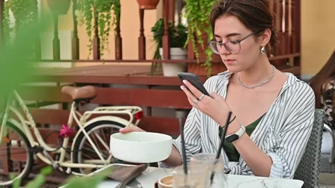 A woman sits at a cafe table, looking at her phone while she has a meal. The sun 스톡 동영상 336778539