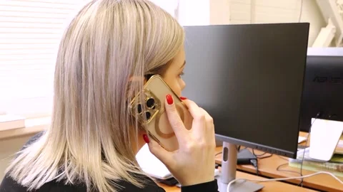 Woman sits at a desk using a computer while talking on a smartphone Stock Footage 335774754