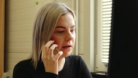 Woman sits at a desk using a computer while talking on a smartphone in a small Stock Footage 336427521