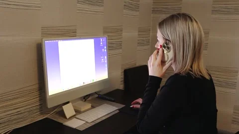 Woman sits at a desk using a computer while talking on a smartphone in a small Stock Footage 336466089