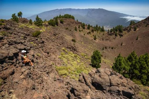Woman sits on the edge of a volcano crater and watches the clouds flow throug Stock Photos
