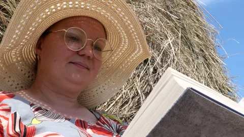 A woman sits in a field next to a haystack and reads a book. Stock Footage 135496786