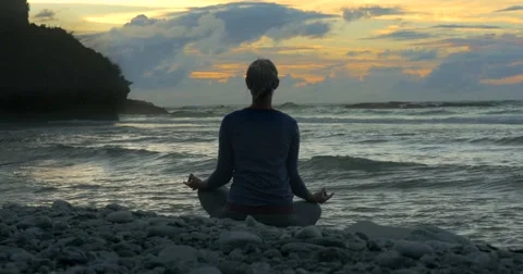 Woman sits in lotus position practicing yoga at sunrise / sunset on beach Stock Footage 59137032