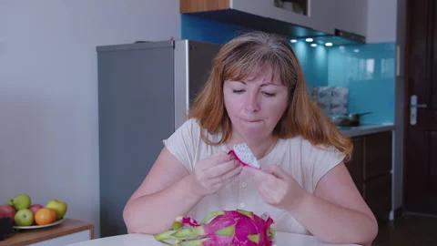 Woman sits at a table in front of a kitchen and eats a dragon fruit Stock Footage 319332734