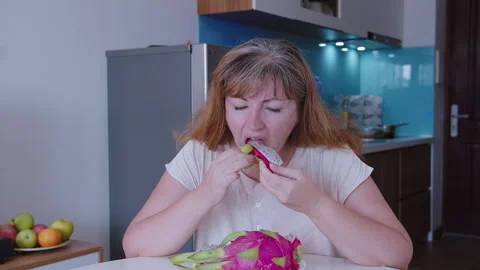 A woman sits at a table in front of a kitchen and eats a dragon fruit Stock Footage 319333645