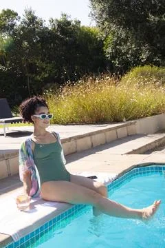 Woman sitting on backyard pool coping, wearing green swimsuit and pastel Foto stock