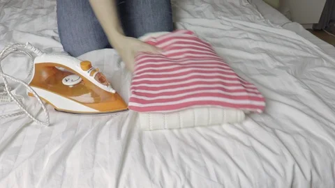 Woman sitting on a bed and putting an iron and a stack of clothes Stock Footage 102621753