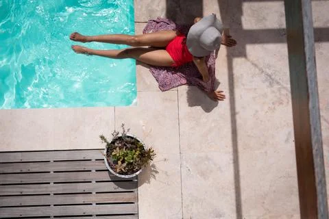 Woman sitting beside rectangular pool on sarong on patio wearing sun hat near Foto stock