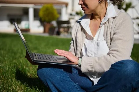 Woman sitting on grass in a yard using a laptop for remote work or study, wea Stock Photos