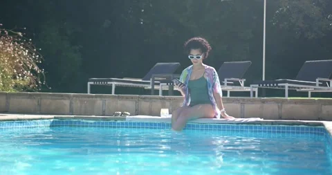 Woman sitting on pool edge, looking at smartphone then kicking water, wearing Stock Footage 344254818