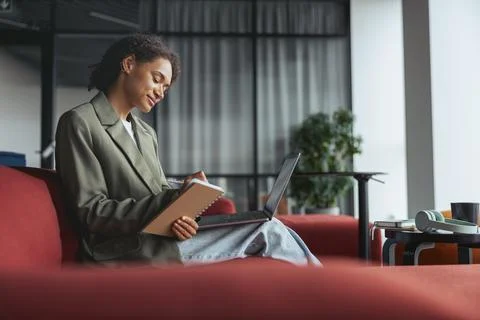 A woman is sitting on a red couch using a laptop and a notebook in nice modern Stock Photos