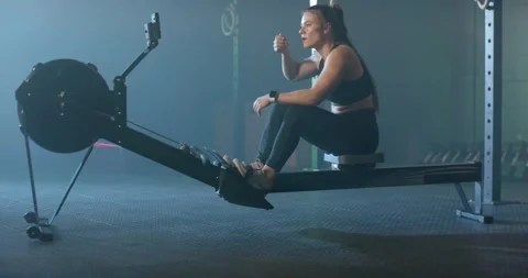 Woman sitting on rower post row wiping forehead, tapping tracker, breathing and Stock Footage 339801644