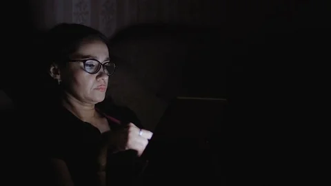 A woman is sitting on a sofa in a dark room and working on a tablet. Stock Footage