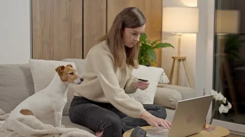 Woman sitting on sofa using laptop and credit card with dog nearby Stock Footage 327306181