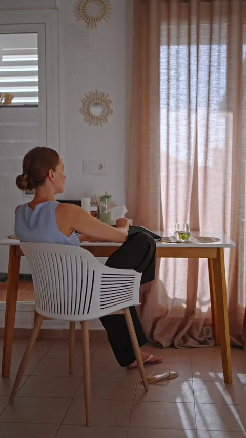 Woman sitting at a table working on a laptop while drinking a beverage at home Stock Footage 341325852