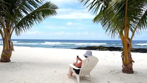 Woman sitting on tropical beach under palm trees using mobile phone device Stock Footage