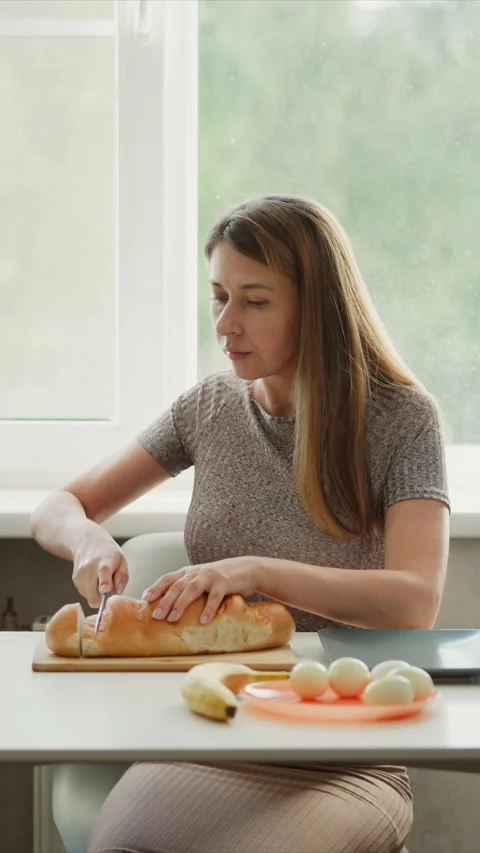 Woman slicing bread with boiled eggs banana on table near window in soft Stock Footage 345980948
