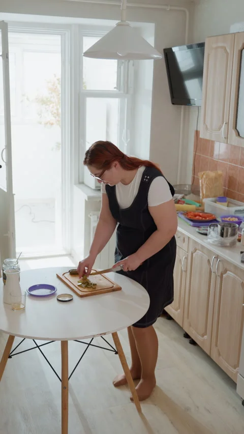 Woman slicing bread at round kitchen table in bright home interior while leaning Stock Footage 343679556