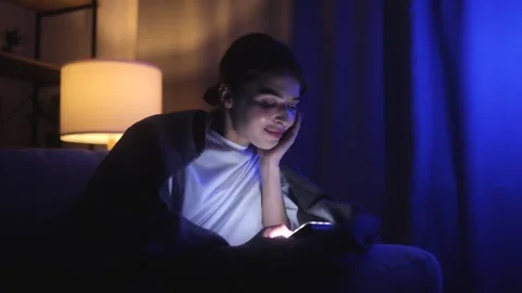 Woman smiling and using smartphone at night under a warm blanket. Cozy home Stock Footage 277887268