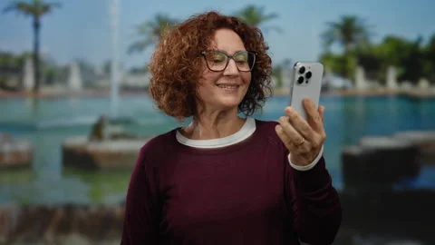 Woman smiling while holding smartphone during a video call in a sunny park .. Stock Footage 312345894
