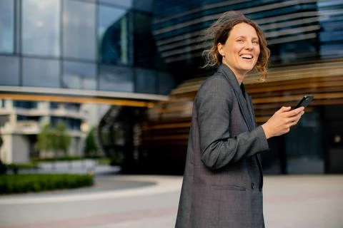 Woman smiling while looking at her phone outside a modern building in the cit 스톡 사진