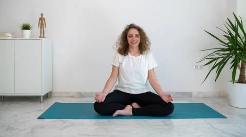 Woman smiling while she does the lotus yoga pose on a blue mat in her home. Stock Photos