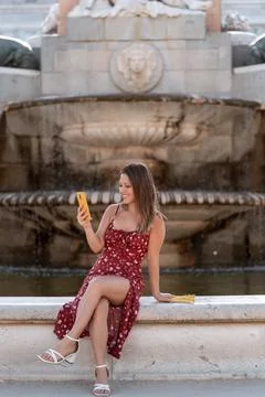 Woman smiling while using her mobile phone sitting outdoors at a water fountain. Foto stock