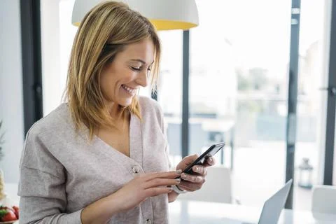 Woman Smiling While Using Smart phone in Bright Kitchen Foto stock