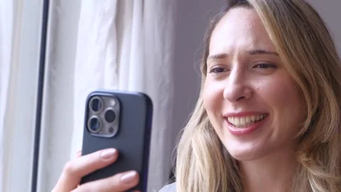 Woman smiling while using a smartphone near a bright window at home. Close-up Stock Footage 328386992