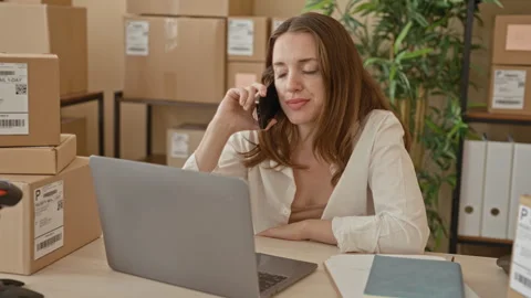 Woman speaking on smartphone while using laptop amid stacked parcels and ba.. Stockbeeldmateriaal 337434546
