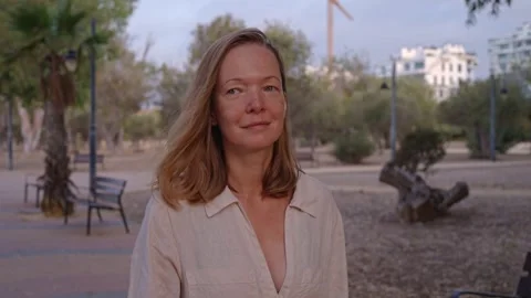 Woman standing in a park looking at the camera with trees and buildings behind Vídeos de archivo 346016832