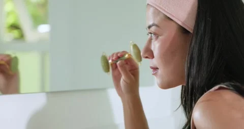 Woman standing at vanity, looking into mirror and rolling green facial roller Stock Footage 335809280