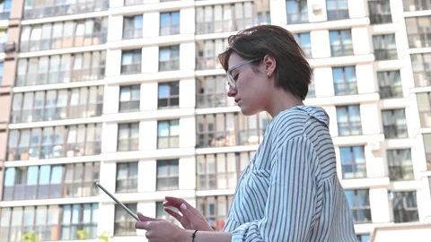 A woman stands outside while using a tablet. She is located in an urban setting Видео 336778218
