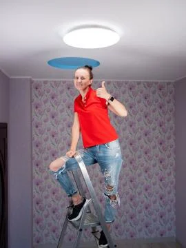 A woman on a stepladder points to an installed led ceiling light and smiles. Stock Photos