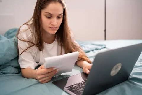 Woman student taking notes while studying using laptop lying on bed at home.. 스톡 사진