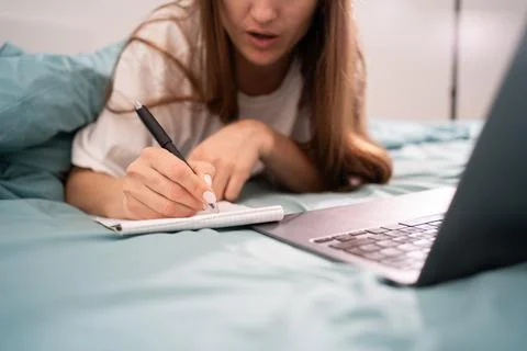 Woman student taking notes while studying using laptop lying on bed at home 스톡 사진