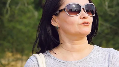 A woman in sunglasses stands against the background of green trees. Video stock 78564371