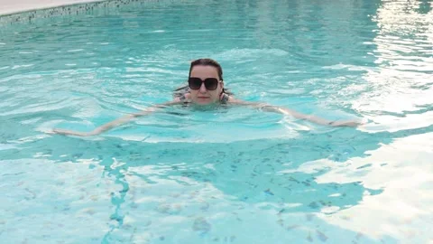 Woman swimming in a clear pool while wearing sunglasses during a sunny day Stock Footage 306413831