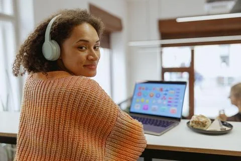 A woman at a table smiles while using a laptop and wearing headphones Stock Photos