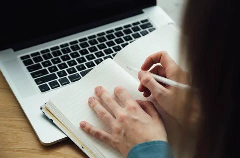A woman takes notes in a notepad using a laptop sitting at the table. Stock Photos