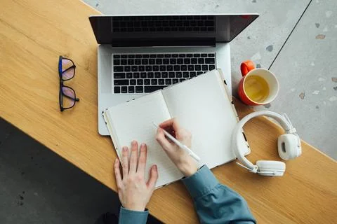 A woman takes notes in a notepad using a laptop sitting at the table, top view. Stock Photos